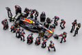 A high-angle shot of a miniature model set depicting an Oracle Red Bull Racing pit stop. The scene features a black, red, and yellow race car surrounded by multiple small pit crew figurines in matching team uniforms, all set against a dark gray reflective surface.