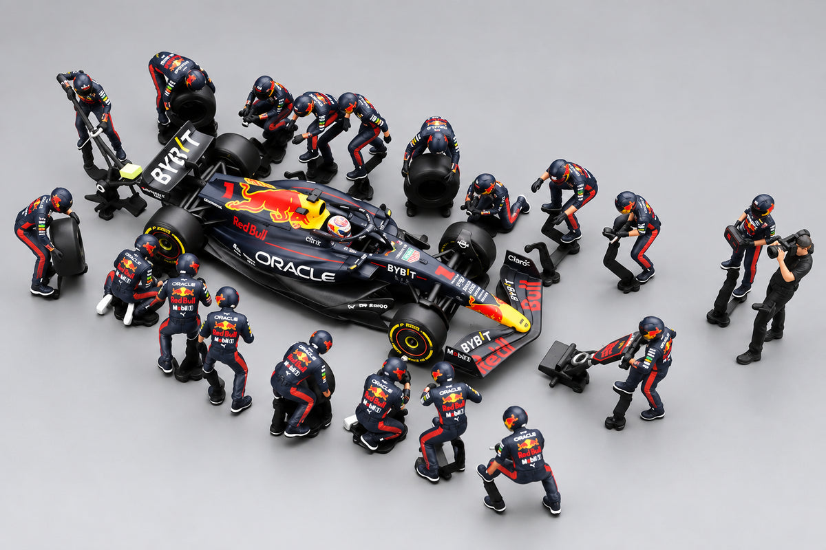 A high-angle shot of a miniature model set depicting an Oracle Red Bull Racing pit stop. The scene features a black, red, and yellow race car surrounded by multiple small pit crew figurines in matching team uniforms, all set against a dark gray reflective surface.